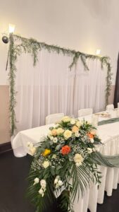 Weiß gedeckter Hochzeitstisch mit Blumengesteck, Vorhänge im Hintergrund.